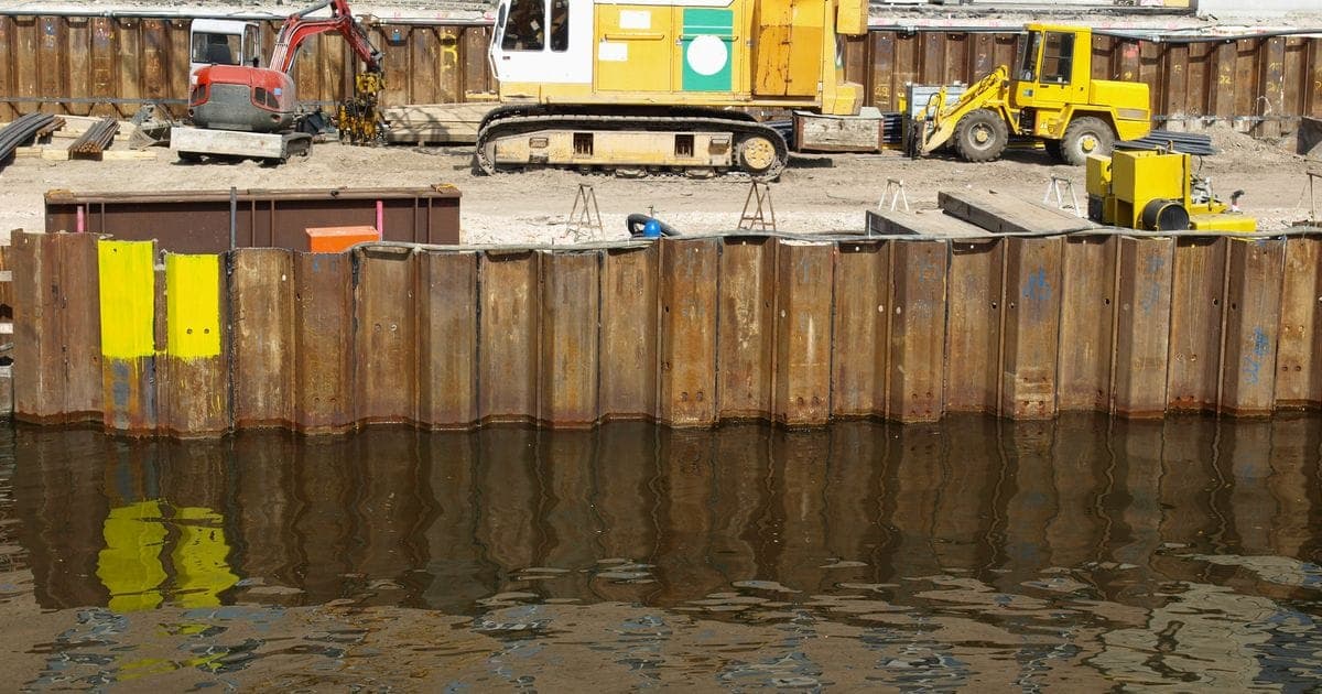Interlocking steel piles driven along the water's edge form a new river wall, with diggers on the site behind.